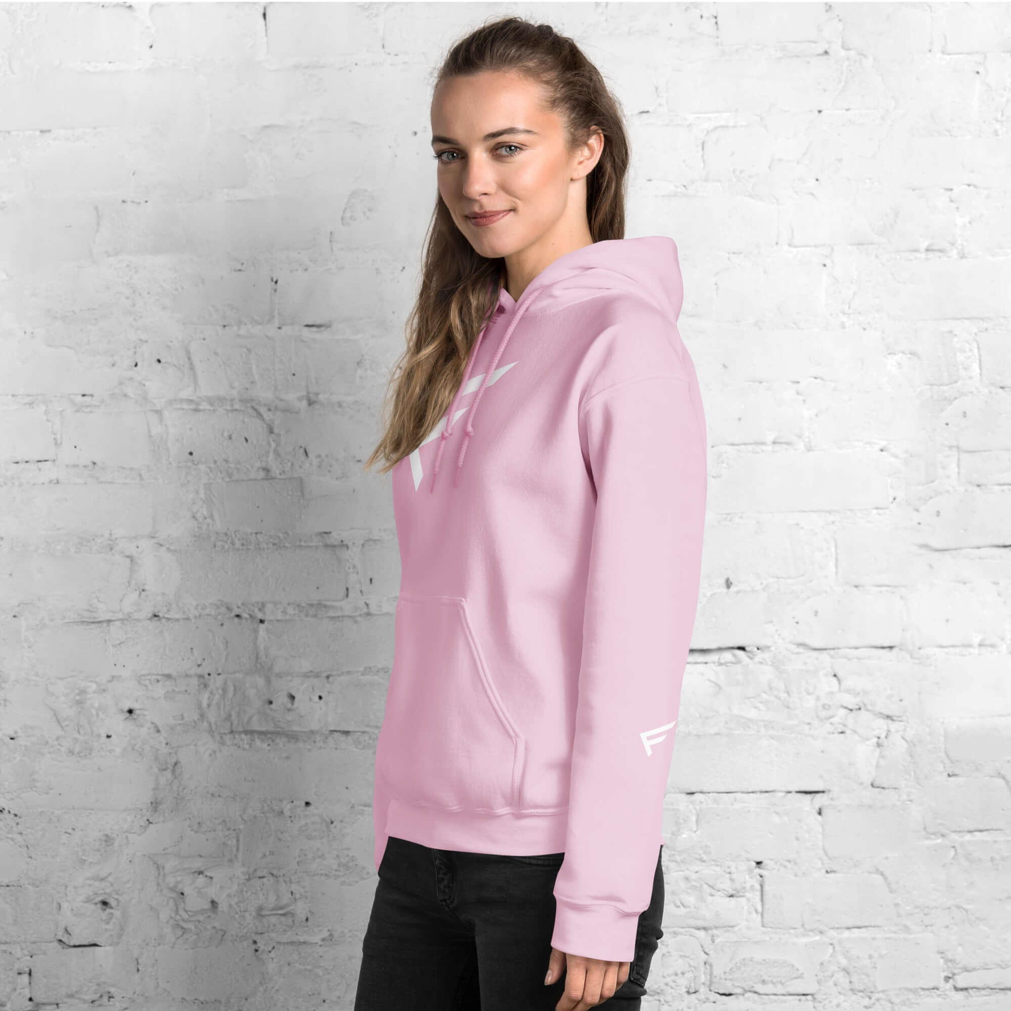 Stylish woman wearing a pink Women's Hoodie featuring signature 'F' emblem against a textured white brick background.